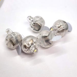 May include: A collection of five clear, spherical LED lights with small, white adhesive tabs. Each light has a small, rounded top and a bright, white LED. The lights are designed for decorative purposes and are arranged against a white background.