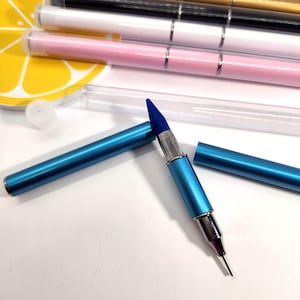 Puede incluir: Una herramienta de nail art azul de doble punta con una punta de silicona azul y una punta de metal. La herramienta es azul metálico con detalles plateados. En el fondo se ven varias otras herramientas de nail art rosas, blancas y negras.