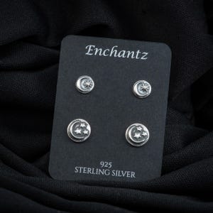 May include: A set of four sterling silver earrings featuring crescent moon and star designs. The earrings are displayed on a black card with the word "Enchantz" and "925 Sterling Silver" printed on it.