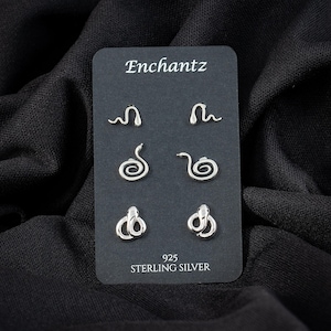 May include: Three pairs of silver-toned snake-shaped stud earrings displayed on a black rectangular card. The card features the text Enchantz at the top and 925 STERLING SILVER at the bottom. Each pair shows a different snake pose.