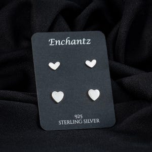 May include: Four sterling silver heart-shaped stud earrings on a black card. The card has the brand name "Enchantz" at the top and "925 STERLING SILVER" at the bottom. The earrings are displayed against a black fabric background.