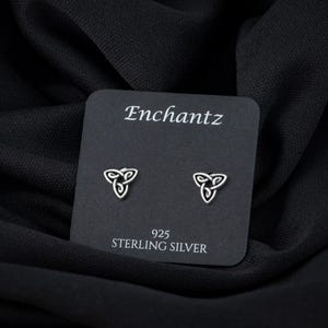 May include: A pair of sterling silver earrings featuring a Celtic knot design. The earrings are presented on a black card with the text "Enchantz" and "925 Sterling Silver" printed on it.