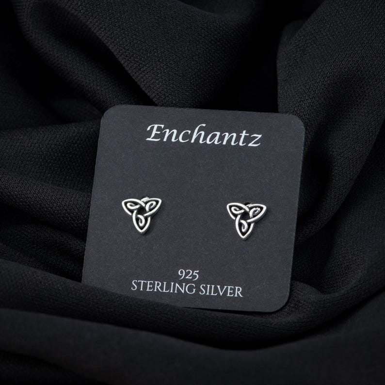 May include: A pair of sterling silver earrings featuring a Celtic knot design. The earrings are presented on a black card with the text "Enchantz" and "925 Sterling Silver" printed on it.
