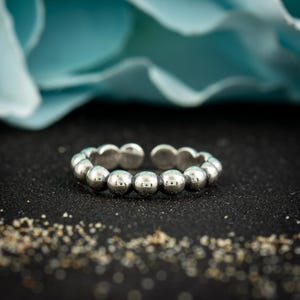 May include: A silver-colored adjustable ring featuring a series of small, round beads. The ring is set against a black background with a blurred blue floral element in the background. The ring's design is simple and elegant.