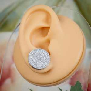 May include: A silver-colored, round stud earring with a surface covered in small, clear stones. The earring is shown on a light-colored ear model, with a soft background.