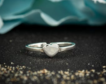 May include: A silver ring with a heart-shaped design. The ring is set against a dark background with a blurred blue flower in the background. The ring's band is thin and delicate.
