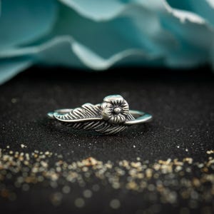 May include: A silver-toned ring featuring a detailed metal feather design that extends along the band, accented with a small, rounded flower-shaped element at the top. The metal surface has a darkened, oxidized finish in the recessed areas to highlight the texture of the leaf veins and the petals of the flower.