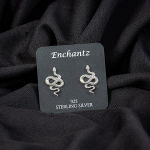 May include: A pair of sterling silver snake stud earrings on a black display card. The card reads "Enchantz" at the top and "925 STERLING SILVER" at the bottom. The earrings are detailed with scales and a curved design.