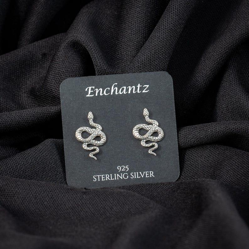 May include: A pair of sterling silver snake stud earrings on a black display card. The card reads "Enchantz" at the top and "925 STERLING SILVER" at the bottom. The earrings are detailed with scales and a curved design.