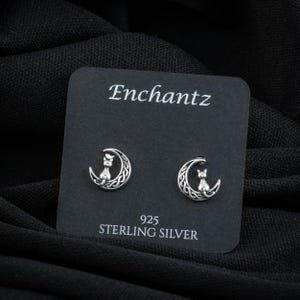 K&ouml;nnte beinhalten: Ein Paar Ohrringe aus Sterlingsilber mit einem Halbmond-Design, auf dem eine Katze sitzt. Die Ohrringe werden auf einer schwarzen Karte mit dem Wort "Enchantz" und "925 Sterling Silver" pr&auml;sentiert.