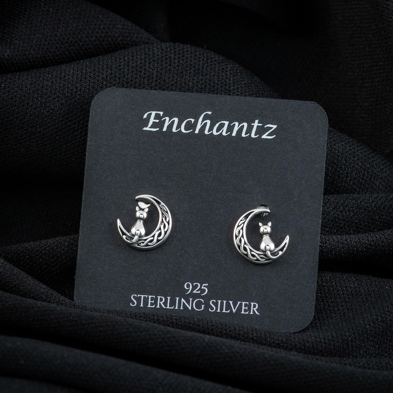 K&ouml;nnte beinhalten: Ein Paar Ohrringe aus Sterlingsilber mit einem Halbmond-Design, auf dem eine Katze sitzt. Die Ohrringe werden auf einer schwarzen Karte mit dem Wort "Enchantz" und "925 Sterling Silver" pr&auml;sentiert.