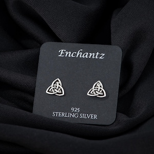 May include: A pair of sterling silver earrings featuring a Celtic knot design. The earrings are displayed on a black card with the brand name "Enchantz" and the text "925 Sterling Silver".