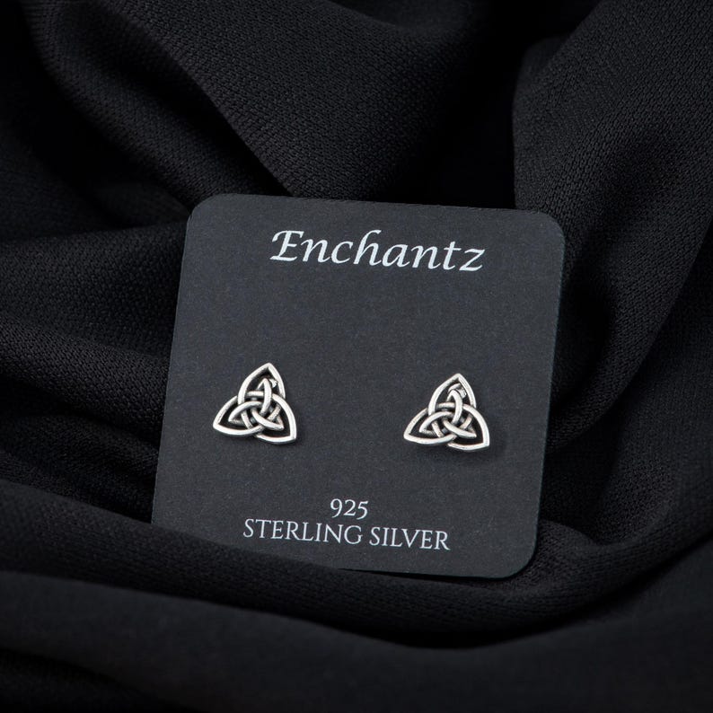 May include: A pair of sterling silver earrings featuring a Celtic knot design. The earrings are displayed on a black card with the brand name "Enchantz" and the text "925 Sterling Silver".