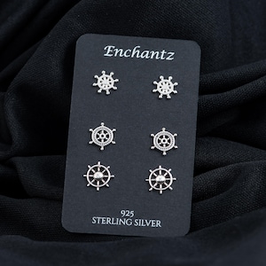 May include: A set of six silver earrings featuring a ship's wheel design. The earrings are displayed on a black card with the word "Enchantz" and "925 Sterling Silver" printed on it.