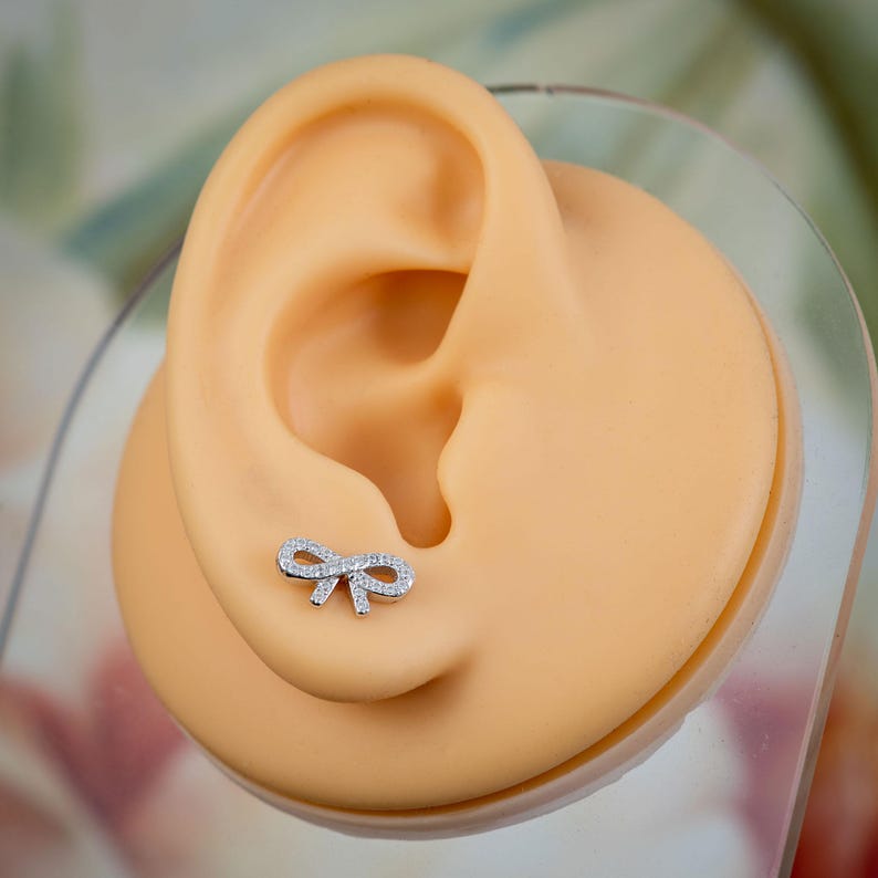 May include: A small, silver bow-shaped earring with clear, sparkling stones. The earring is displayed on a tan ear model. The bow design is delicate and detailed, with the stones adding a touch of elegance. The background is blurred, with hints of green and white.