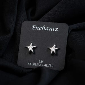 May include: A pair of silver star-shaped stud earrings with a central clear stone, mounted on a black display card labeled Enchantz and 925 Sterling Silver. The earrings feature a polished metallic finish and a five-pointed star design.