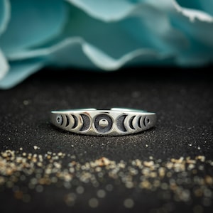 May include: A silver ring featuring a crescent moon phase design. The ring has a series of crescent moon shapes and a central full moon. The ring is set against a black background with a blurred blue backdrop.