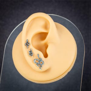 May include: Three silver-colored snake earrings displayed on a tan ear model. The earrings are different sizes and designs, with intricate detailing. The ear model is set against a dark gray background, showcasing the jewelry.