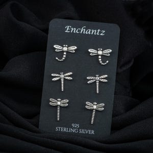 May include: Six sterling silver dragonfly stud earrings displayed on a black card. The card is labeled "Enchantz" and "925 Sterling Silver". The earrings are detailed with wings and a slender body. The background is a dark, textured fabric.
