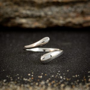 May include: A silver-colored ring with a modern, open design. The ring features two teardrop-shaped ends that curve around the finger. The ring is set against a dark, textured surface.