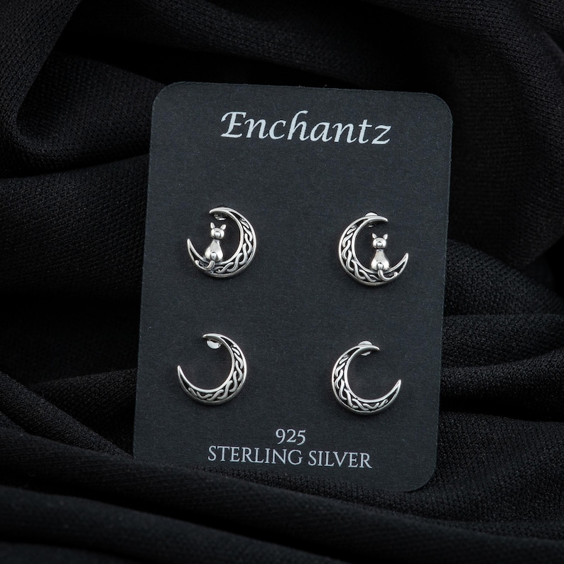 K&ouml;nnte beinhalten: Ein Set mit vier silbernen Ohrringen, die einen auf einem Halbmond sitzenden Katzen zeigen. Die Ohrringe werden auf einer schwarzen Karte mit dem Text "Enchantz 925 Sterling Silver" pr&auml;sentiert.