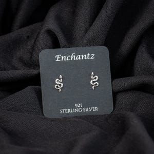 May include: A pair of sterling silver snake stud earrings on a black card. The card reads "Enchantz" at the top and "925 STERLING SILVER" at the bottom. The earrings are small, silver, and shaped like snakes.