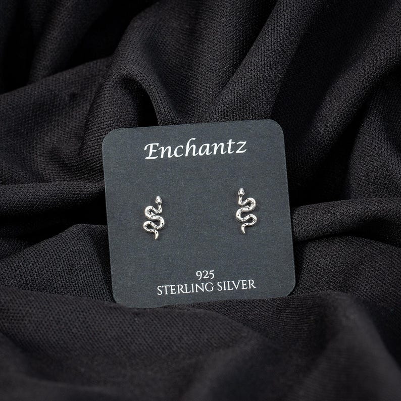 May include: A pair of sterling silver snake stud earrings on a black card. The card reads "Enchantz" at the top and "925 STERLING SILVER" at the bottom. The earrings are small, silver, and shaped like snakes.