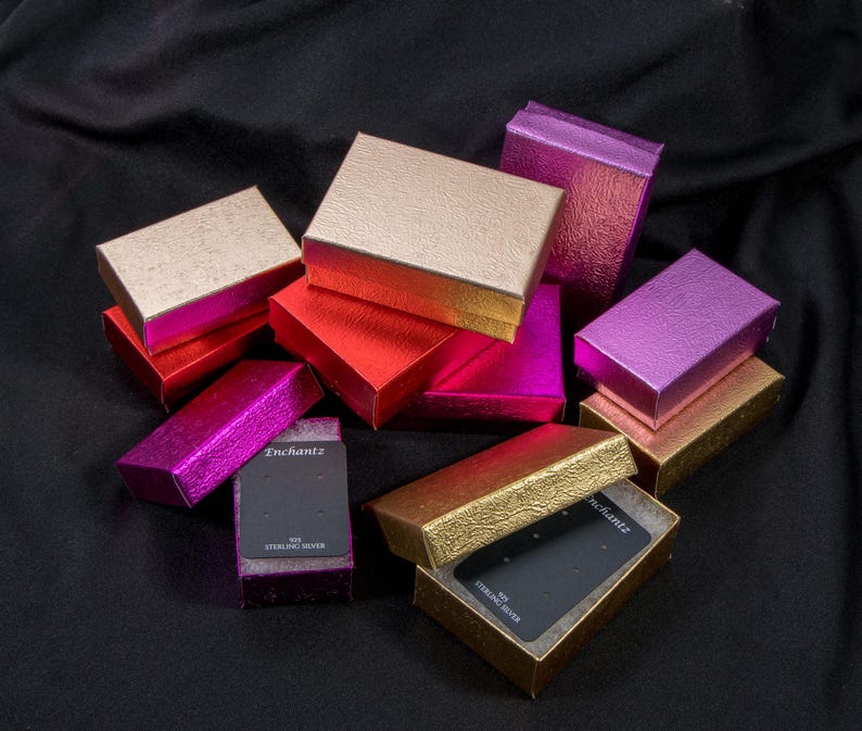 May include: An assortment of small, colourful boxes with a textured, metallic finish. Some boxes are stacked on top of each other. The boxes are made of cardboard and have a black interior. The boxes are labelled "Enchantz" and "925 Sterling Silver".