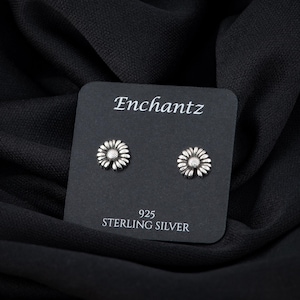 May include: A pair of silver daisy stud earrings on a black card with the text "Enchantz 925 Sterling Silver".