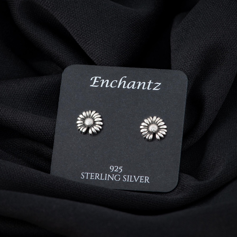 May include: A pair of silver daisy stud earrings on a black card with the text "Enchantz 925 Sterling Silver".