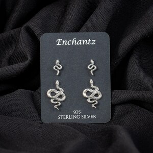 May include: Silver snake earrings on a black card with the text "Enchantz" and "925 STERLING SILVER". The earrings are in the shape of snakes, with two pairs of different sizes.