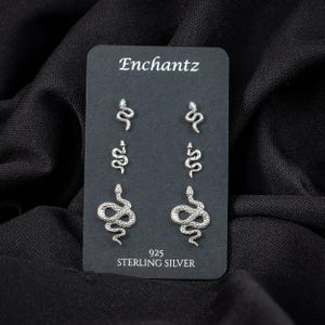 May include: A black card displays six sterling silver snake stud earrings. The card reads "Enchantz" at the top and "925 STERLING SILVER" at the bottom. The earrings are in three different sizes and designs.