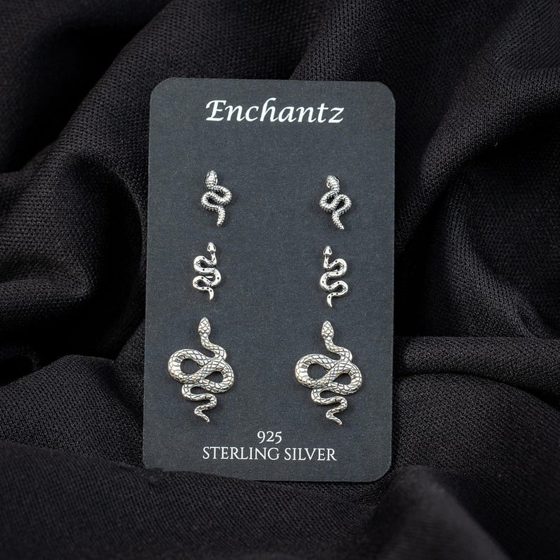 May include: A black card displays six sterling silver snake stud earrings. The card reads "Enchantz" at the top and "925 STERLING SILVER" at the bottom. The earrings are in three different sizes and designs.