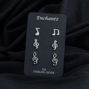 May include: A set of six sterling silver earrings featuring music note and treble clef designs. The earrings are displayed on a black card with the brand name "Enchantz" and the text "925 Sterling Silver".