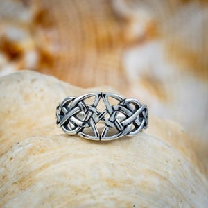 May include: A silver-colored ring with a Celtic knot design. The ring features interwoven patterns and a central star-like shape. The ring is displayed on a light-colored, textured surface, possibly a seashell.