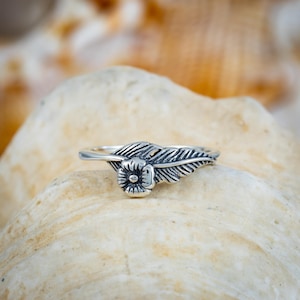 May include: A silver ring featuring a floral design with a feather detail. The ring is set against a neutral background, highlighting the intricate details of the jewelry. The ring's design includes a small flower and a detailed feather.