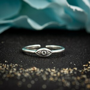 May include: A silver-toned open ring featuring a central eye-shaped motif. The band is smooth and polished, while the eye design includes detailed etched lines and a small circular center, creating a distinct focal point on the jewelry piece.