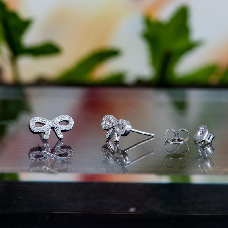 May include: A pair of silver bow-shaped stud earrings, encrusted with small, clear gemstones. The earrings are displayed on a reflective surface, with the earring backs and posts visible. The background is blurred with green and white hues.