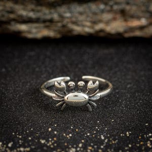 May include: A silver-colored ring featuring a detailed crab design. The crab has prominent claws, eyes, and a textured body. The ring is set against a dark, textured background, highlighting the jewelry's intricate details.