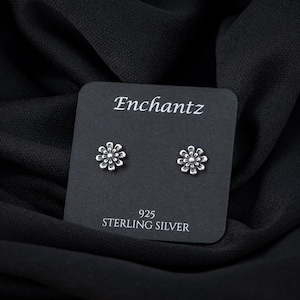 May include: A pair of silver flower stud earrings on a black card with the text "Enchantz 925 Sterling Silver".