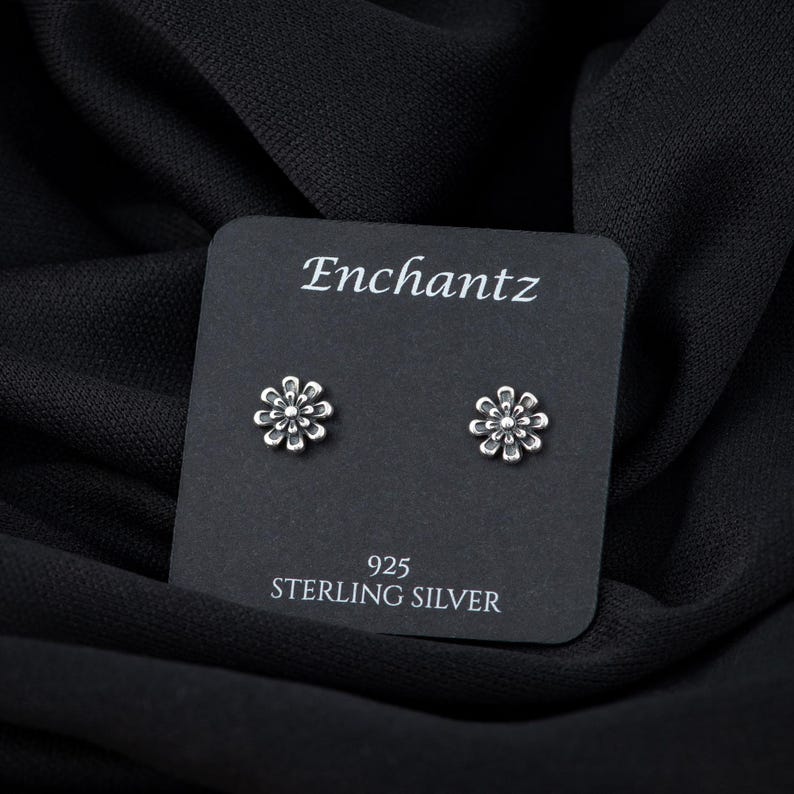 May include: A pair of silver flower stud earrings on a black card with the text "Enchantz 925 Sterling Silver".