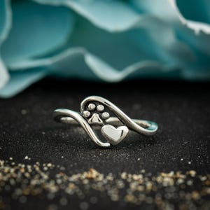 May include: A silver ring featuring a paw print and a heart design. The ring has a flowing, curved shape and is set against a dark background with a blurred blue backdrop.