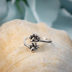 May include: A silver-colored ring featuring two paw print designs. The ring is wrapped around the finger with the paw prints facing opposite directions. The ring is set against a neutral background.