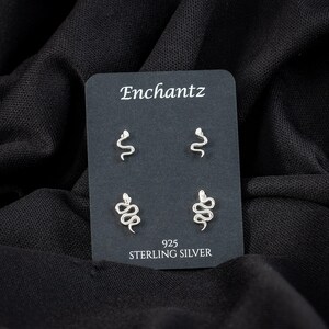 May include: Two pairs of silver snake-shaped stud earrings displayed on a black rectangular card. The card features the text Enchantz at the top and 925 STERLING SILVER at the bottom. The earrings are arranged in two rows with a smaller pair above.