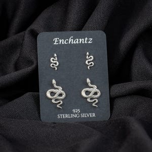 May include: A black card displays four silver snake earrings. The card reads "Enchantz" at the top and "925 Sterling Silver" at the bottom. The earrings are in two different sizes, with two small and two larger snake designs.