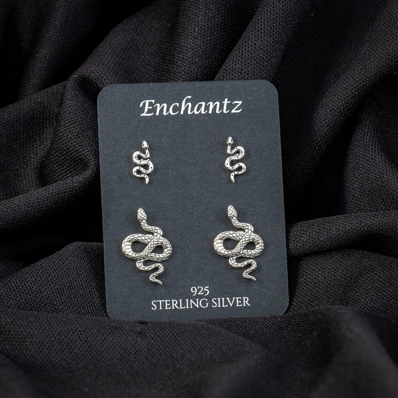 May include: A black card displays four silver snake earrings. The card reads "Enchantz" at the top and "925 Sterling Silver" at the bottom. The earrings are in two different sizes, with two small and two larger snake designs.