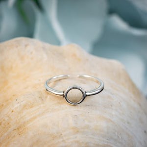 May include: A silver-colored ring with an open circle design at the center. The ring is set on a light-colored, textured surface, with a soft, blurred background. The ring's band is thin and adjustable.