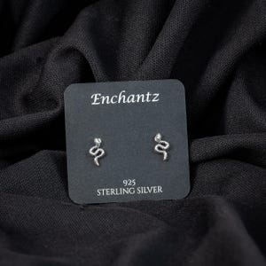 May include: A pair of sterling silver snake stud earrings on a black card with the text "Enchantz" and "925 STERLING SILVER". The earrings are small, silver, and shaped like snakes.