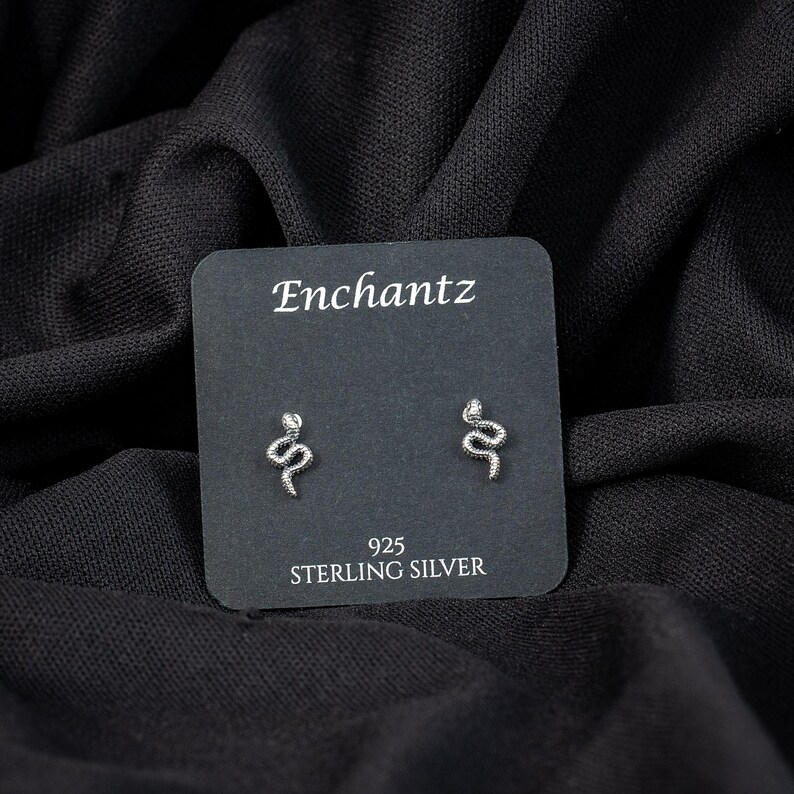May include: A pair of sterling silver snake stud earrings on a black card with the text "Enchantz" and "925 STERLING SILVER". The earrings are small, silver, and shaped like snakes.