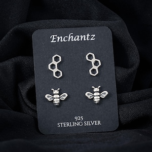 May include: A black card displays two pairs of silver earrings. One pair is a honeycomb design, and the other is a bee. The card reads "Enchantz" at the top and "925 STERLING SILVER" at the bottom.